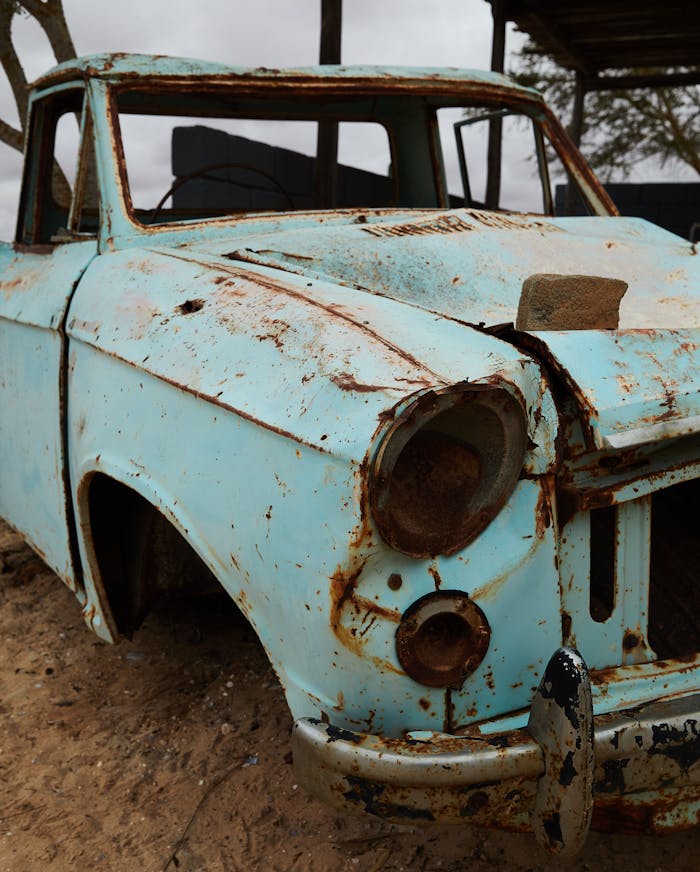 A rusted and abandoned vintage car sits decaying in a sandy outdoor setting.